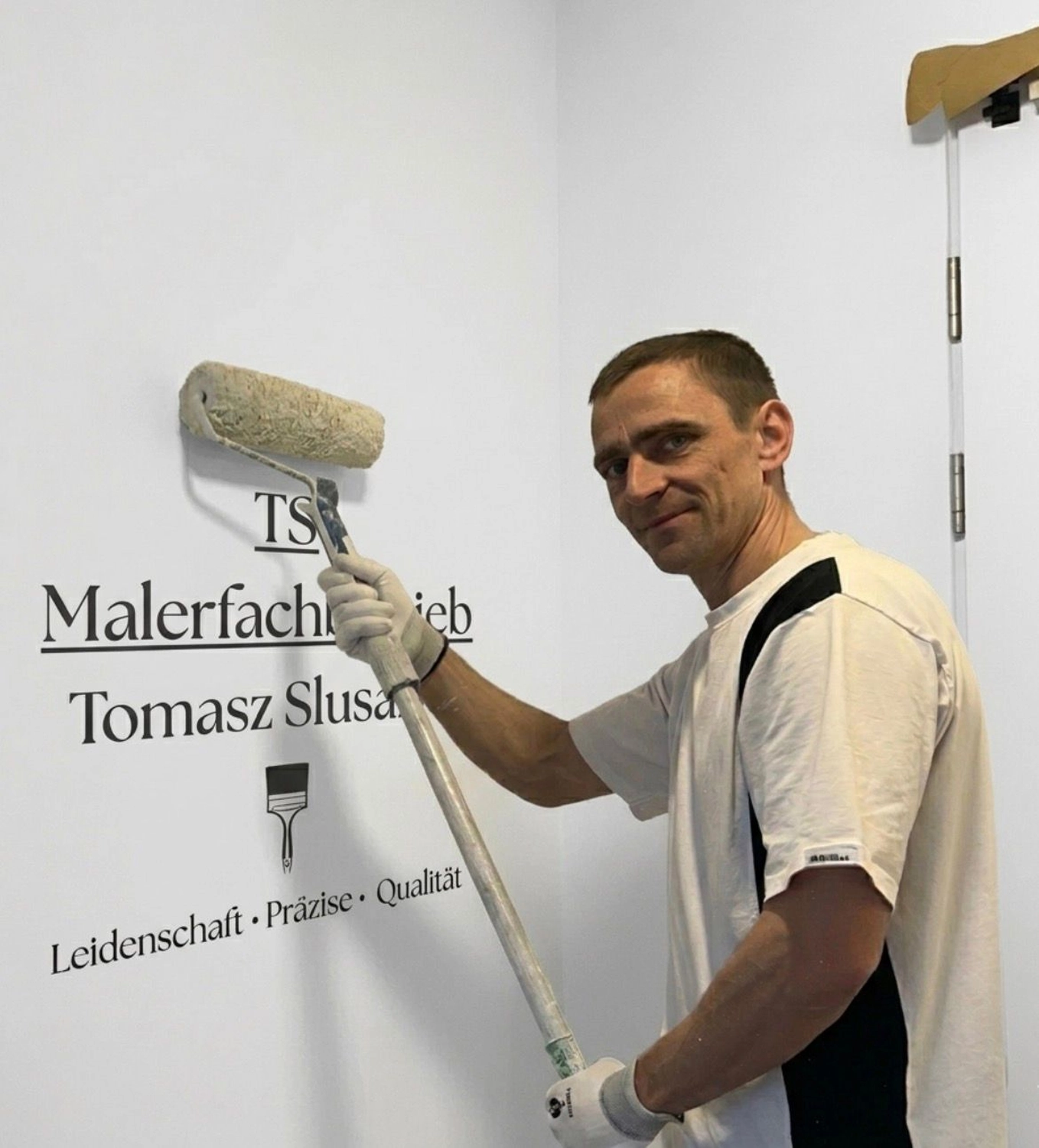 TS Malerfachbetrieb - Maler und Lackierer - Tomasz Slusarek mit Farbroller in der Hand und Firmenlogo an der Wand